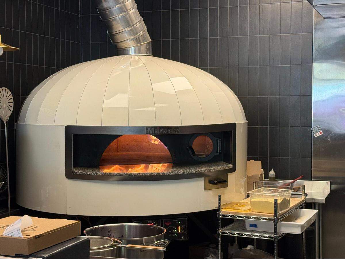 A straight-on, eye-level shot of a large, modern, dome-shaped pizza oven. The oven is cream-colored and has a wide, dark opening, revealing a roaring fire and glowing wood embers inside. A large, silver ventilation pipe extends from the top of the dome. The oven is set against a wall of sleek, black, rectangular tiles. To the right, a small metal prep cart holds containers, including one filled with yellow cornmeal.