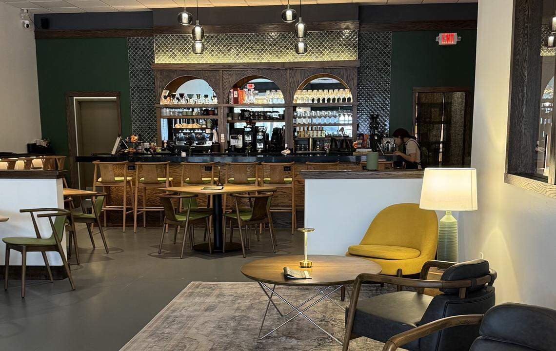 A wide, eye-level shot of a restaurant interior showing a lounge area with a yellow armchair in the foreground. In the mid-ground, small tables with olive-green chairs lead to the main bar area, which is framed by dark wood arches and dark green tile.