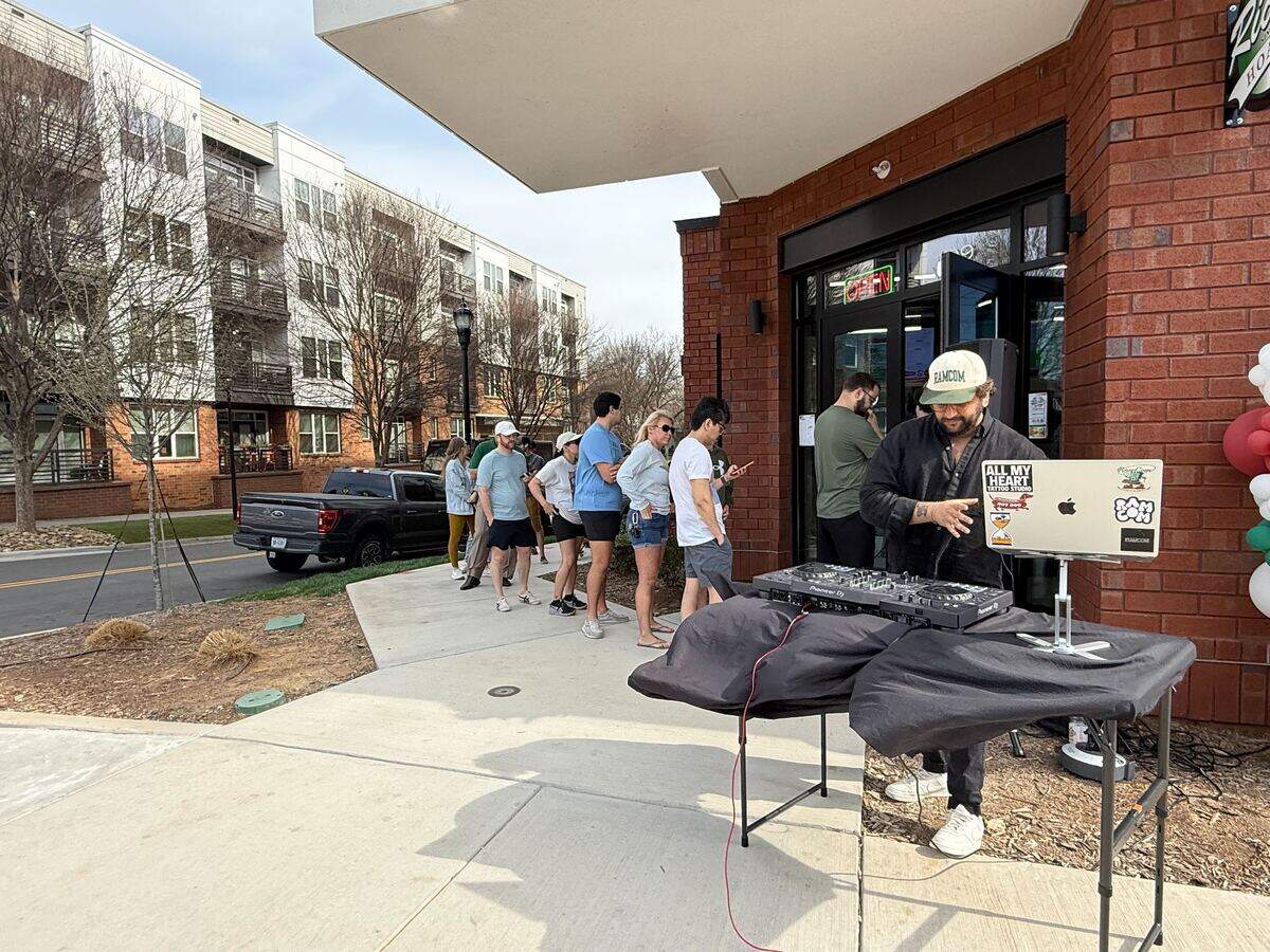 A DJ wearing a black shirt and a “ZAMCOM” baseball cap performs on a sidewalk outside the entrance of “Ricci’s Hoagies.” The DJ setup includes a Pioneer controller and a laptop adorned with various stickers, positioned on a table covered with a black cloth. In the background, a line of customers waits to enter the brick building, and modern apartment complexes are visible across the street.