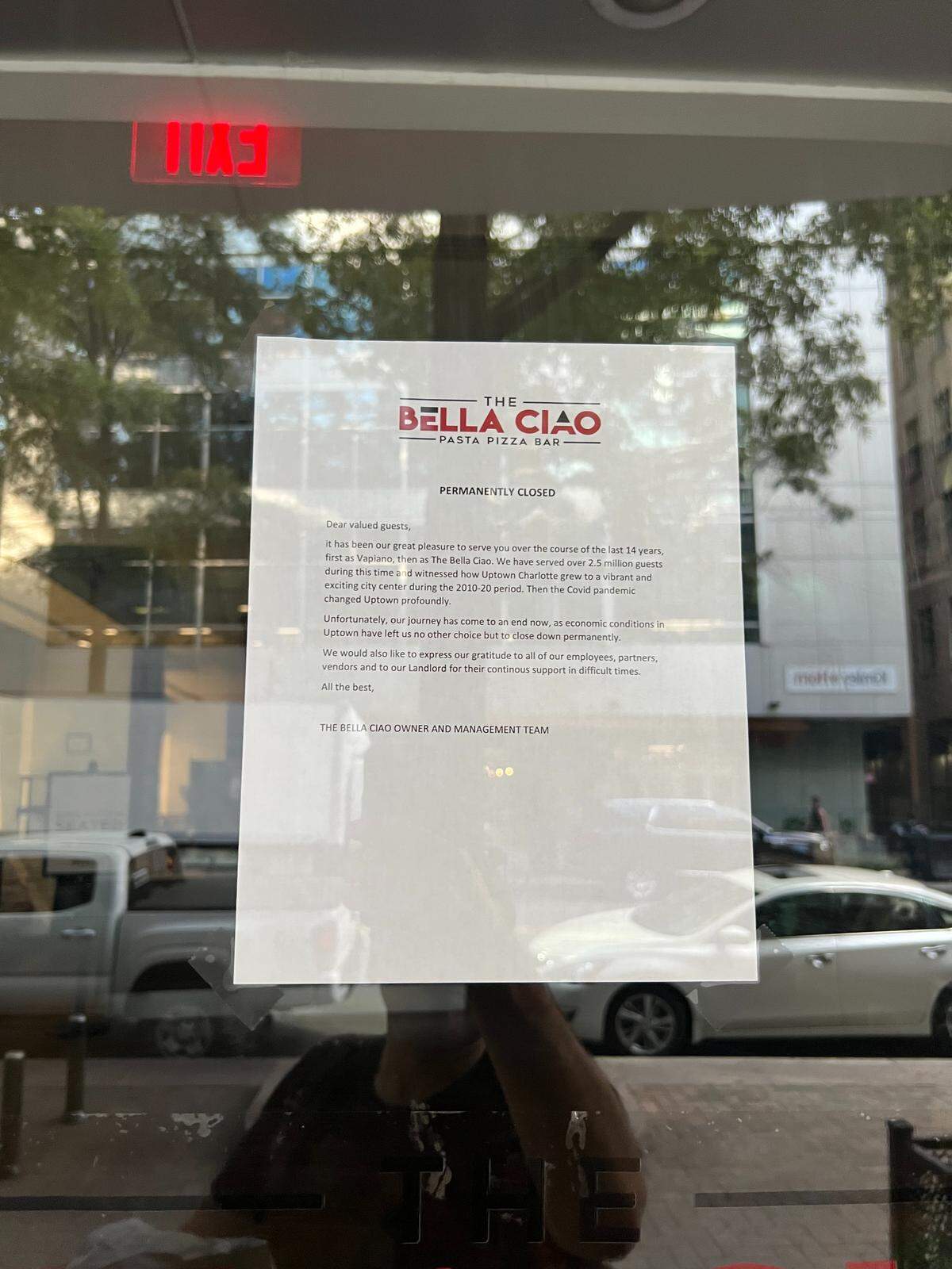 A “Permanently Closed” sign from “The Bella Ciao Pasta Pizza Bar” is taped to the glass door of the restaurant. The letter on the sign thanks guests for their patronage, and reflections of the street are visible on the glass.