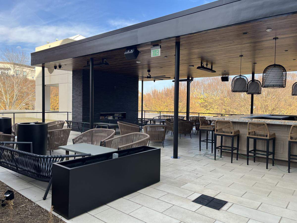 A wide, eye-level shot of a modern outdoor patio, partially covered by a black roof with a wood plank ceiling. The terrace features an outdoor bar area and lounge seating furnished with contemporary rope-style chairs and stools. The patio overlooks a dense line of trees displaying brilliant autumn foliage.