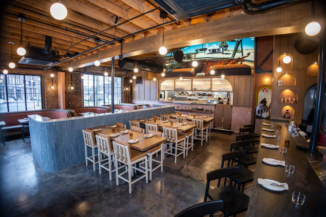 A focused view of the dining area highlighting tall, light-wood tables with woven-back chairs. In the background, a large graphic mural of a person in a vintage car overlooking a desert landscape sits above a pass-through window into the kitchen.