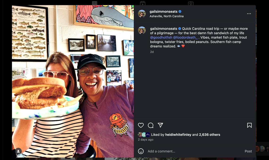 A screenshot of an Instagram post from user gailsimmonseats (Gail Simmons). The photo within the post shows two smiling women taking a selfie in a casual restaurant. The woman on the left holds up a large fried fish sandwich on a paper plate, and the woman on the right smiles broadly. The post’s caption describes a trip to Asheville, North Carolina for “the best damn fish sandwich of my life” at the restaurant Good Hot Fish.