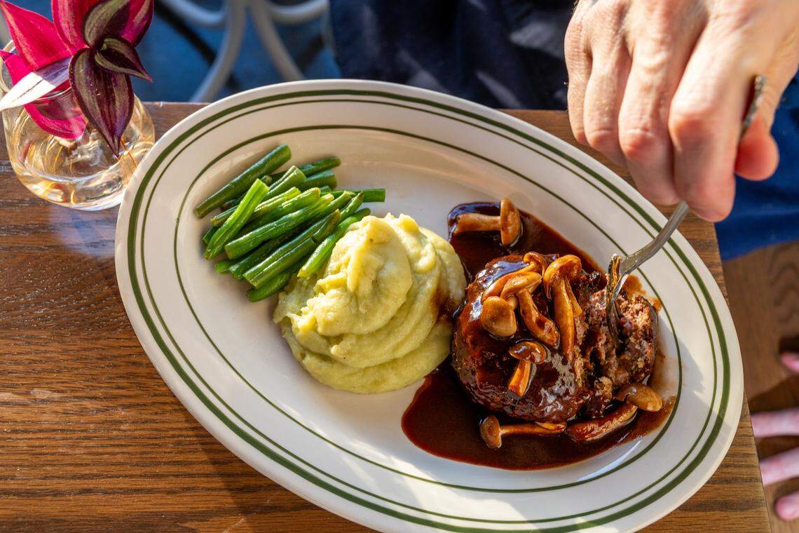 An overhead view of a Salisbury steak smothered in dark gravy and topped with whole sautéed mushrooms. It is served with a large scoop of mashed potatoes and a side of bright green beans on an oval diner plate.