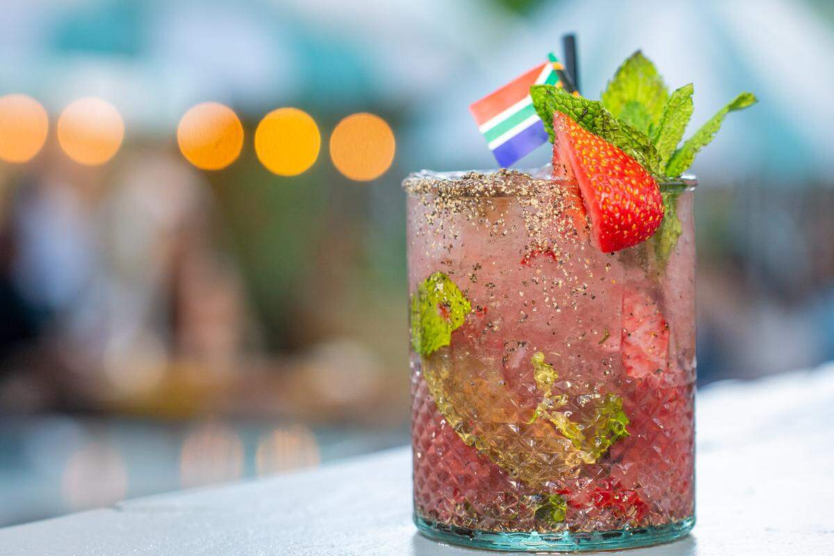 A close-up shot focuses on a cold, textured glass of a bright pink cocktail with a sugared or spiced rim, sitting on a bar or table. The drink is garnished with a fresh strawberry slice, mint sprigs, and a tiny South African flag on a black pick.