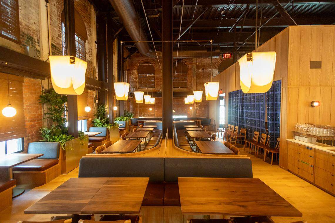 An interior, wide-angle shot of a large, high-ceilinged dining room with a symmetric layout. The center of the room features two long, dark grey banquettes arranged back-to-back, separated by a light wood divider that runs down the middle of the space.