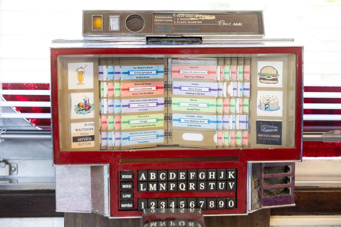 A close-up shot of a vintage Rowe AMI tabletop jukebox selector box at a diner. The retro machine features a red frame, a silver alphanumeric keypad at the bottom, and a glass display showing colorful song title strips. Visible artists include Buffalo Springfield, Carrie Underwood, and Pink Floyd. The sides of the display are decorated with small illustrations of a milkshake, a burger with fries, and a slice of cake.