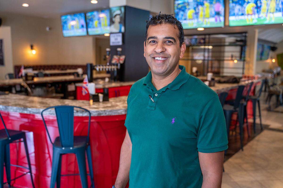 A medium, eye-level shot of a smiling restaurant owner in a dark green polo shirt, standing inside the Big Ben Pub. The pub’s distinctive red U-shaped bar, blue barstools, and a row of televisions displaying a soccer game are visible in the blurred background.
