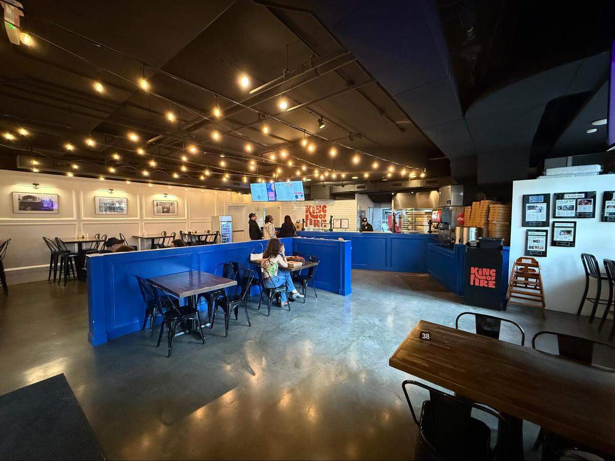 A wide interior shot of the “King of Fire” restaurant. The space features polished concrete floors, blue partitions, and dark ceilings draped with warm string lights. A few customers are seated at tables, and the pizza prep area is visible in the back under bright lights.