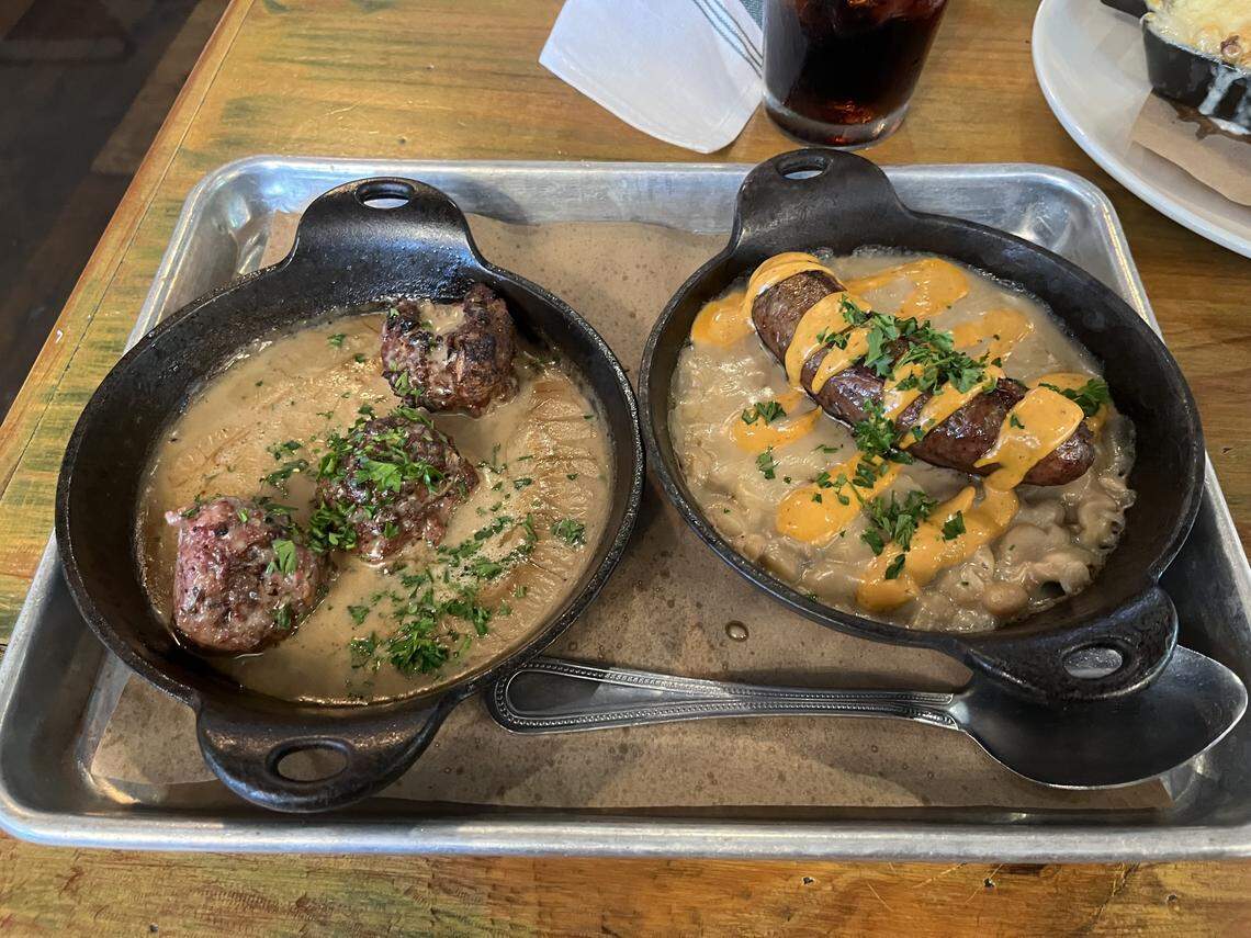 A close-up view of two small, cast-iron skillets served on a metal tray, resting on brown parchment paper. The skillet on the left contains two large meatballs submerged in a pale brown sauce, garnished with parsley. The skillet on the right holds a sausage link over a bed of white beans in a sauce, drizzled with a bright orange sauce and garnished with parsley. A spoon rests on the tray below the skillets.