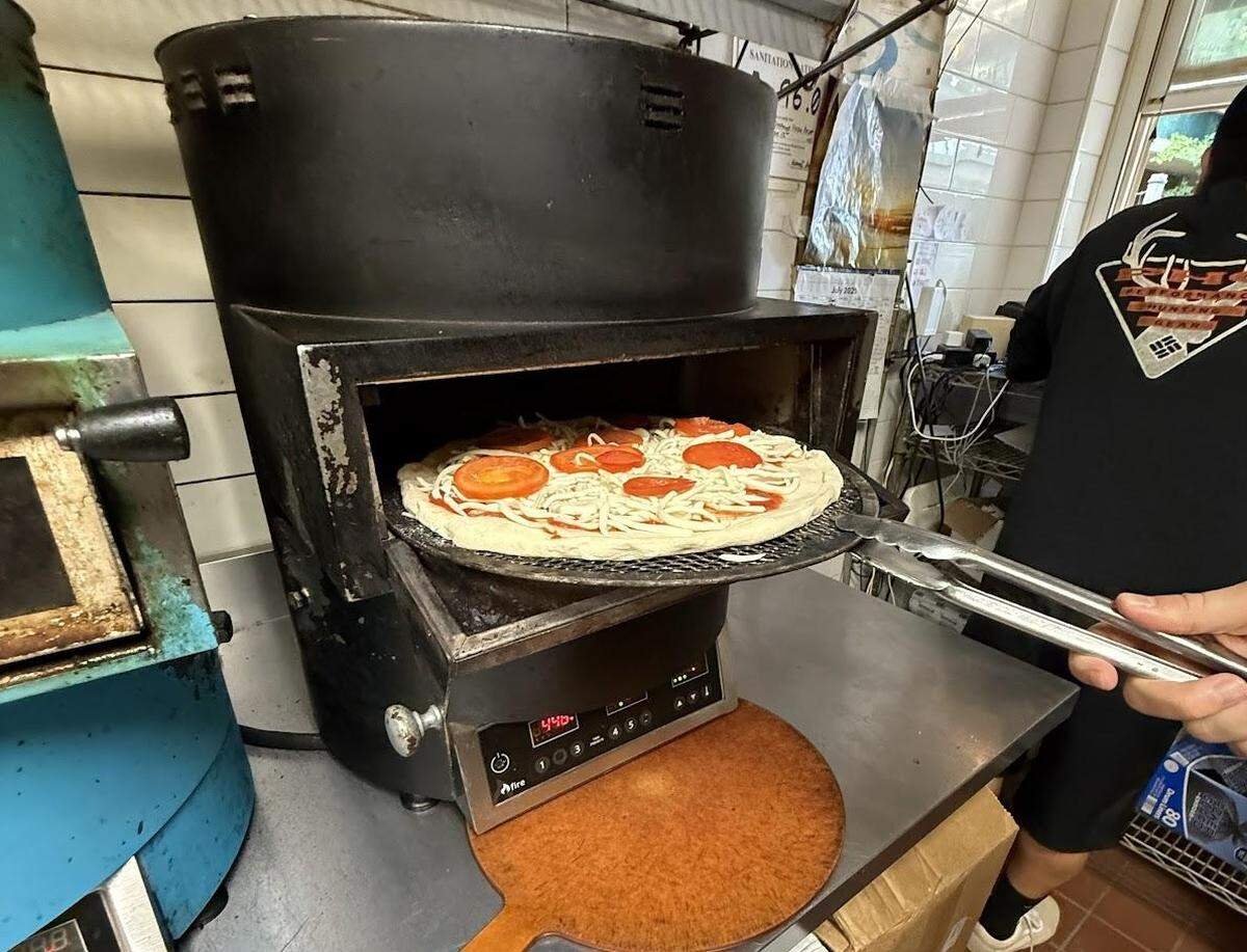 A close-up action shot from a pizzeria kitchen shows a cook using long tongs to place an uncooked tomato and cheese pizza into a specialized black oven. The pizza slides off a round mesh screen into the oven’s opening. The oven’s digital control panel is visible below.