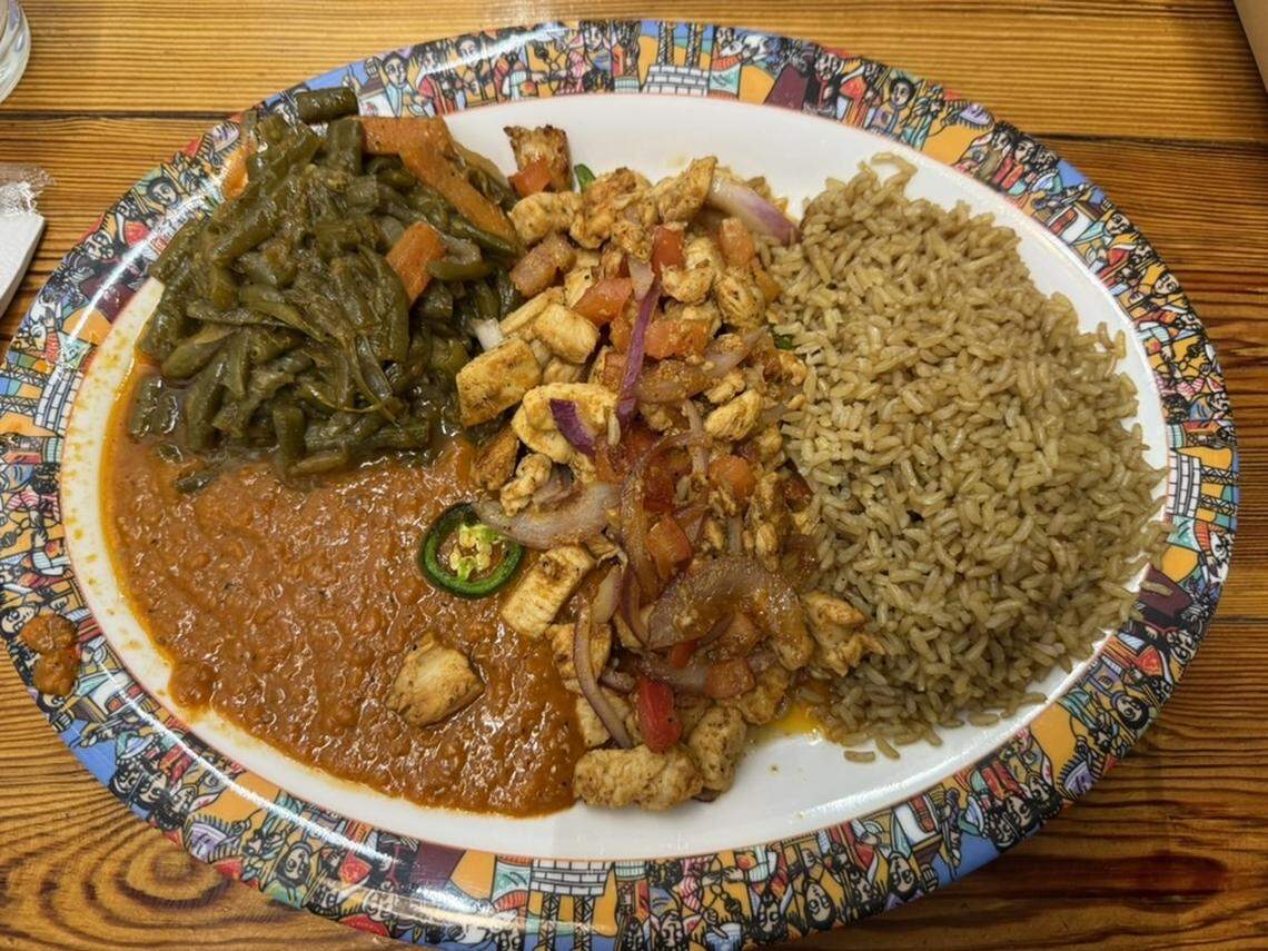 A top-down view of a large white plate with a highly decorative, multi-colored rim featuring illustrations of people. The plate is filled with several distinct portions of food: a pile of seasoned brown rice on the right, a heap of sautéed chicken with onions and peppers in the center, a serving of cooked green beans and carrots on the top left, and a generous portion of thick, reddish-brown lentil stew (misir wot) on the bottom left, topped with a single slice of fresh green jalapeño. The plate sits on a rustic wooden table.