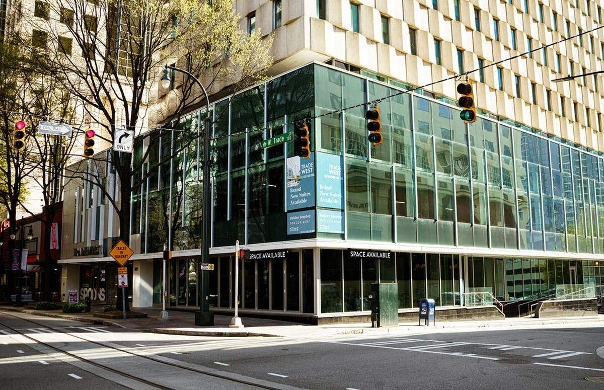 An exterior view of a modern commercial building with extensive glass paneling at the street corner, contrasting with the lighter, older façade of the high-rise structure behind it. The corner is marked by a traffic light and street signs, and a banner on the glass advertises “Trade West: Brand New Suites Available.”