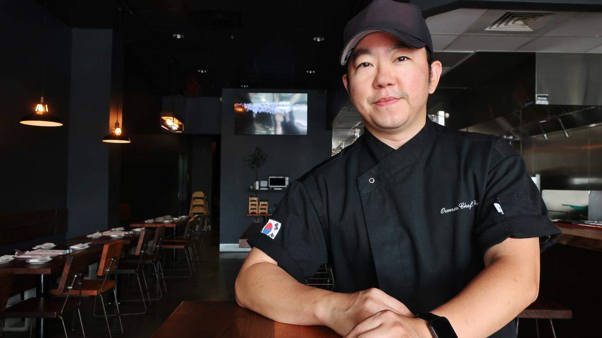 A close-up portrait of a chef seated at a wooden table inside his restaurant wearing a black chef’s coat with a South Korean flag patch on the sleeve and a black baseball cap. The restaurateur is looking toward the camera with a pleasant expression and hands clasped.
