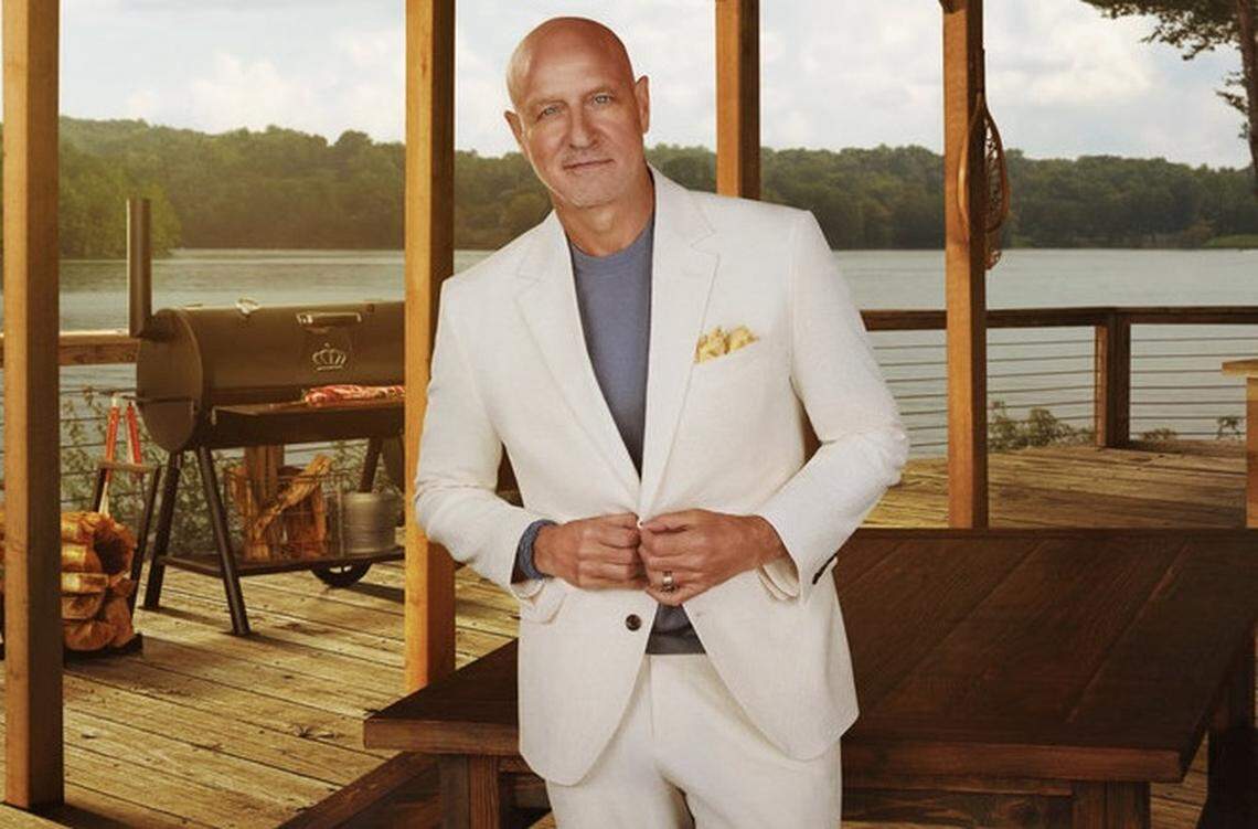 A person is standing at the center of the frame, looking directly at the camera with a slight, confident smile and dressed in a white linen suit jacket over a blue-gray t-shirt, featuring a yellow patterned pocket square. The person is positioned on a rustic wooden deck under a wooden pergola with exposed pillars.