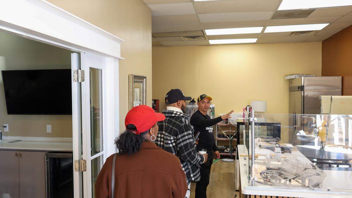 A person in a black long-sleeve shirt and baseball cap gestures toward the interior of a brightly lit commercial space. Two people, one wearing a red hat and the other a plaid jacket, stand in the foreground looking into the room, which contains a white counter and large stainless steel appliances.