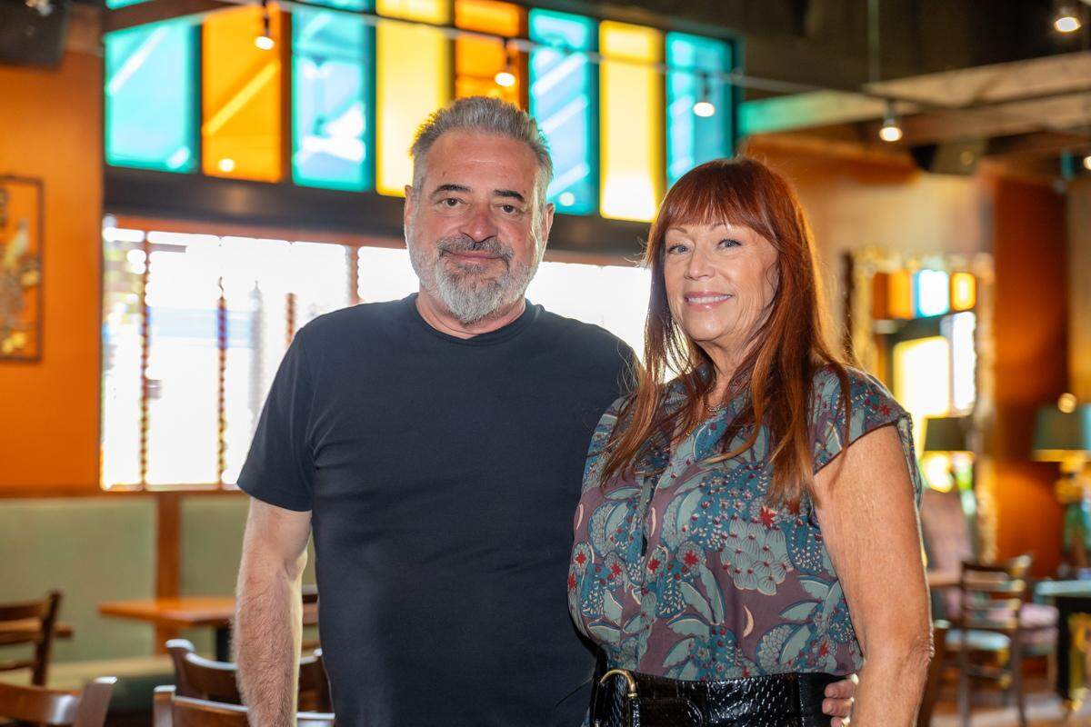 Soul Gastrolounge owners Andy and Lesa Kastanas smiling for the camera inside their restaurant. Andy has a gray beard and a black t-shirt, and Lesa has reddish-brown hair and a patterned blouse. The restaurant’s colorful windows are visible in the background.