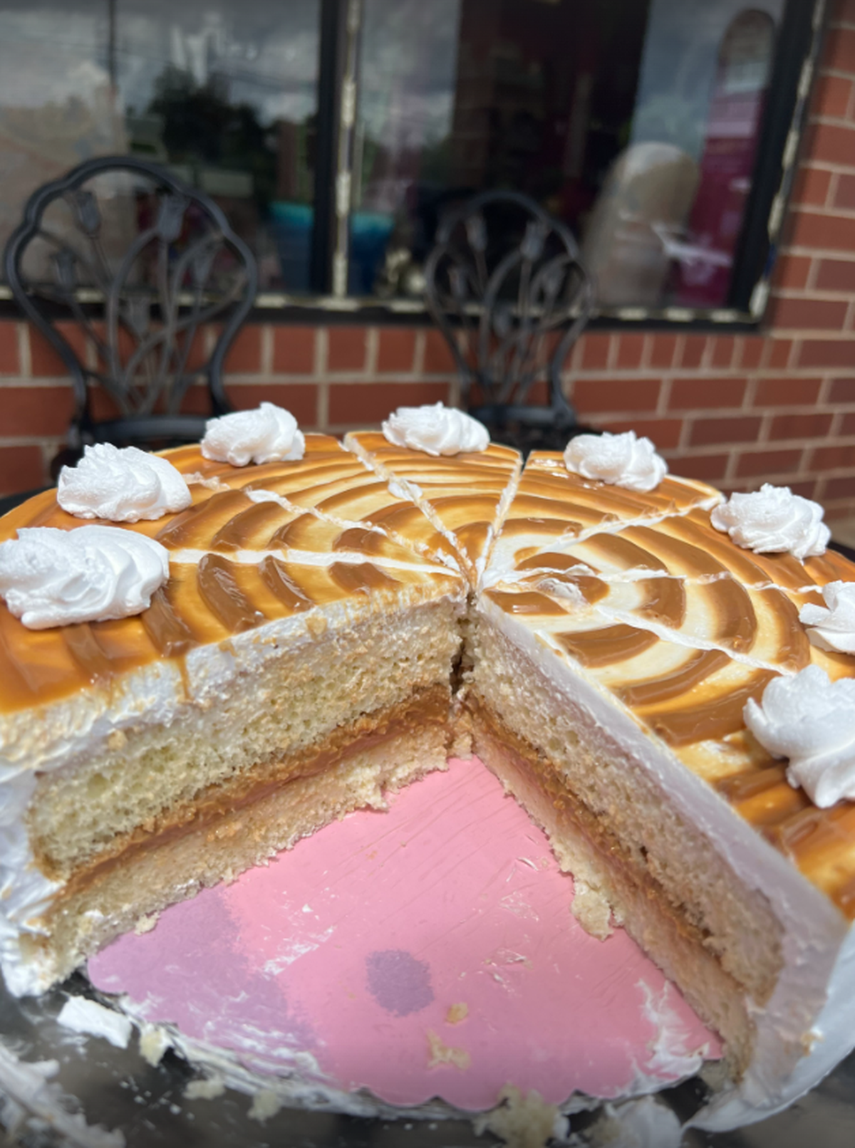 A delicious-looking tres leches cake is shown with a slice already cut out. The cake has two layers of light-colored cake with a caramel or dulce de leche filling between them. The top of the cake is covered in a caramel sauce with white icing piped in a spiral pattern, and there are several dollops of whipped cream around the edge. In the blurred background, two black metal chairs are visible.