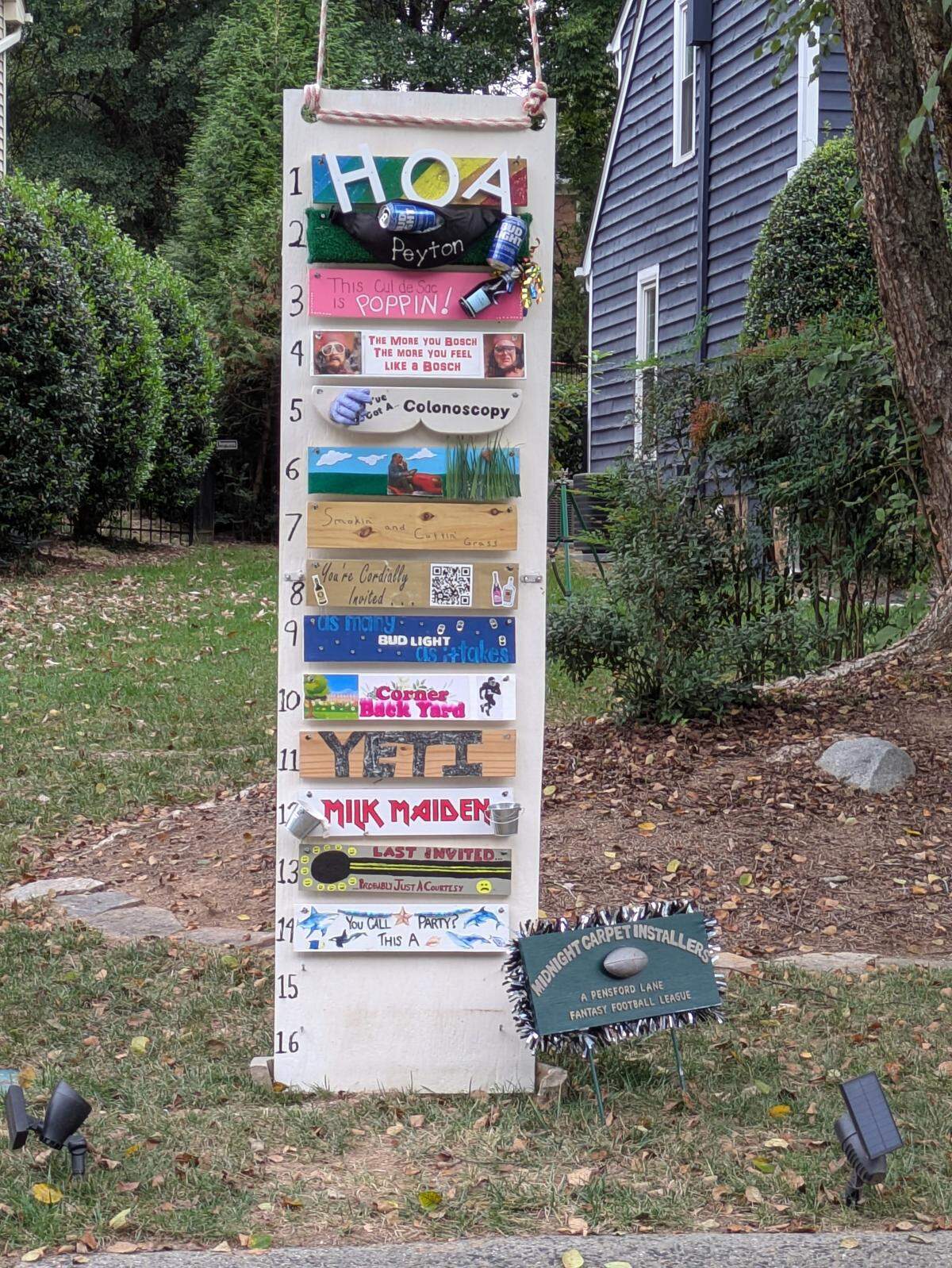 A full shot of a tall, white, wooden, homemade sign standing in the front yard of a house. The sign serves as a humorous fantasy football league ranking, with numbers 1 through 16 listed vertically. Various decorated and themed wooden planks are attached horizontally, each representing a different team or rank, featuring jokes and names like “HOA,” “Colonoscopy,” “Corner Back Yard,” and “LAST INVITED.” A smaller green sign at the bottom identifies the league as the “MIDNIGHT CARPET INSTALLERS” and “A FENSTERD LANE FANTASY FOOTBALL LEAGUE.”