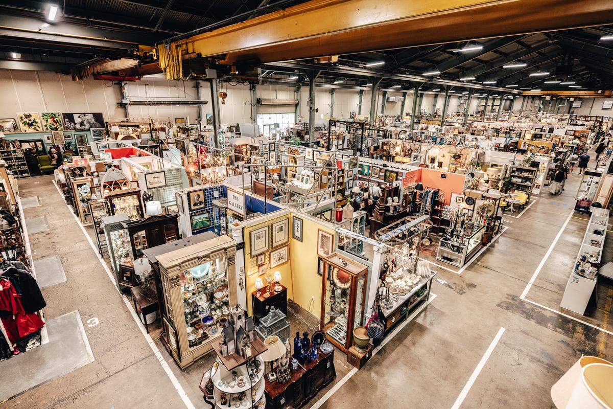 From a high vantage point, the camera looks down on a massive indoor antique mall. The industrial-style building features a yellow overhead crane and a concrete floor divided into a grid of stalls. Each stall is densely filled with vintage furniture, curio cabinets, paintings, and countless other second-hand goods.