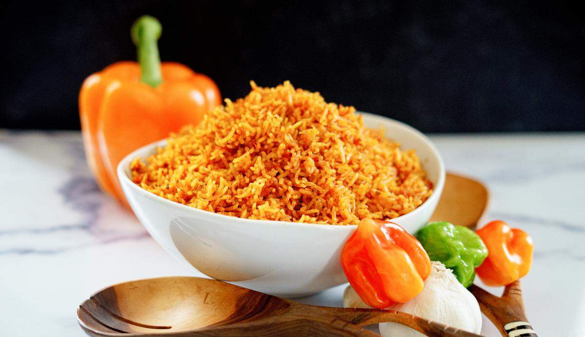 A white bowl overflowing with bright orange Jollof rice is centered on a white marble-patterned surface. In the background, a large, whole orange bell pepper is visible. In the foreground, surrounding the base of the bowl, there are three wooden spoons, two bulbs of garlic, and three small, colorful peppers: one bright green and two orange (likely Habanero or Scotch Bonnet). The background is a blurred black surface.