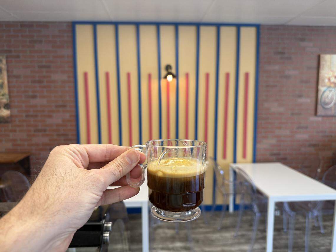 A hand holds a clear glass mug containing a dark coffee drink, possibly an Affogato, with a visible scoop of creamy gelato melting into the hot espresso. The background features the shop’s signature striped wall and clean, white tables, creating a cozy and modern aesthetic.