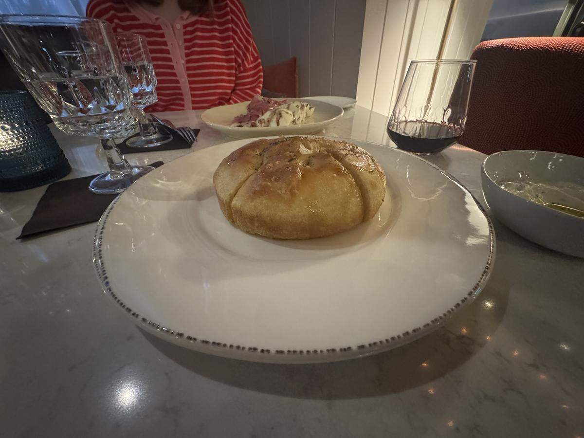 A close-up shot of a golden-brown, round loaf of focaccia bread, sliced into six wedges, served on a large white plate with a dark-speckled rim. The table is white marble, and a person in a red-and-white striped shirt sits in the background. Crystal water glasses and a stemless glass of red wine are visible next to the plate.