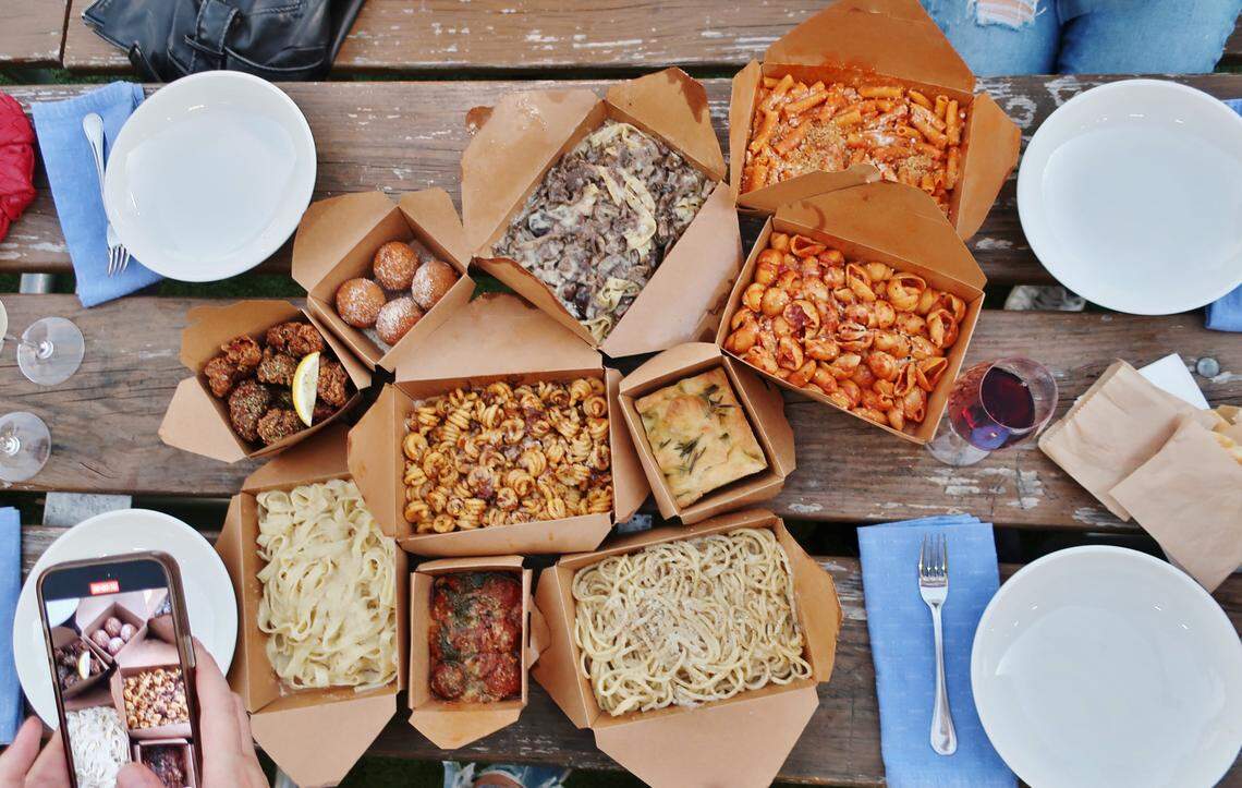 An overhead view of a wooden picnic table covered with multiple open brown cardboard takeout containers filled with various Italian dishes, including different types of pasta, meatballs, fried dough balls, and focaccia. Around the containers are white plates, blue cloth napkins, silverware, and a hand holding a phone taking a photo of the food.