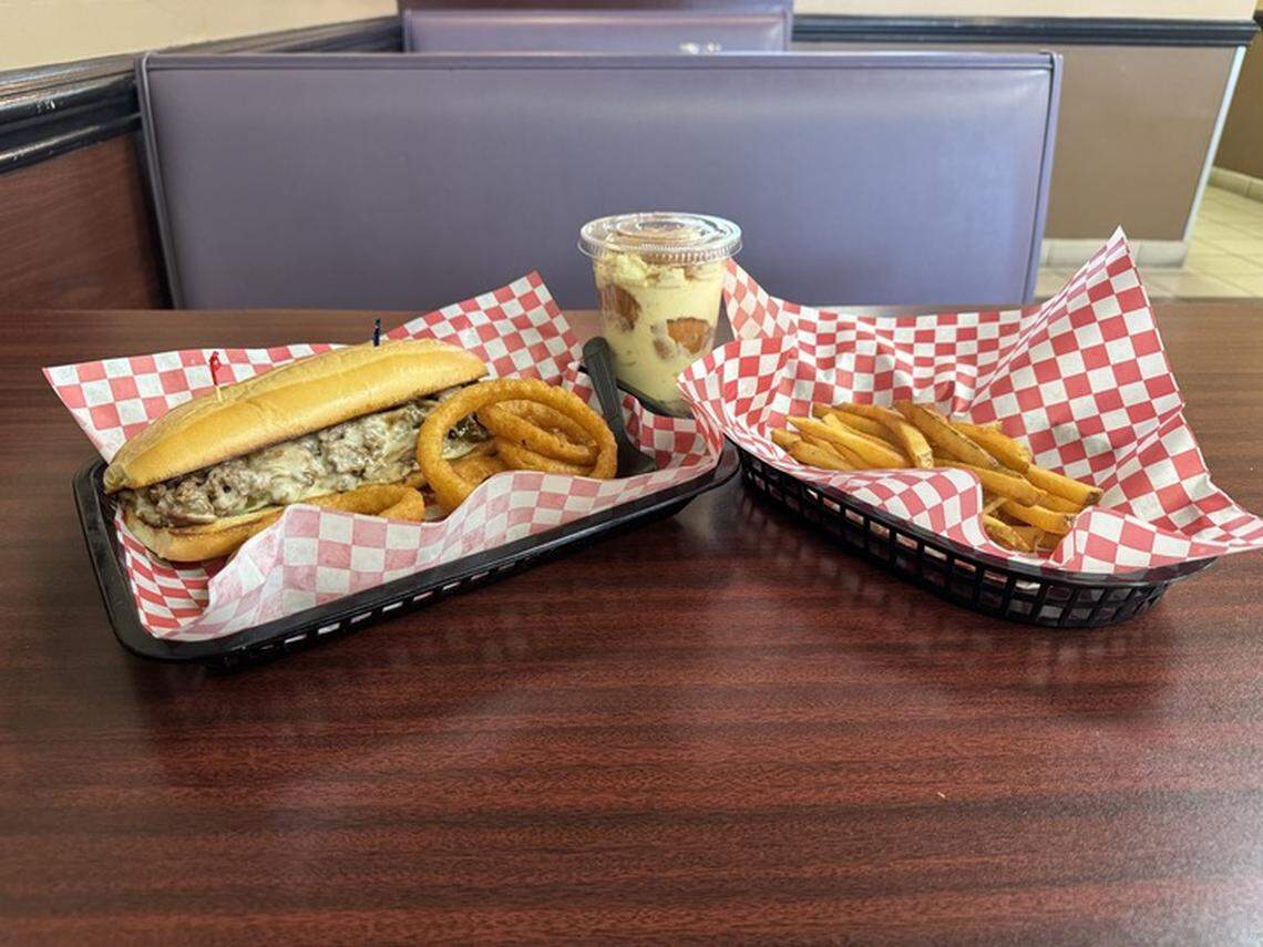 A cheesesteak sub with a side of onion rings, a separate container of french fries, and a cup of what appears to be banana pudding. The food is served on two black plastic baskets lined with red and white checkered paper. The meal is on a brown wooden table in a booth with a purple vinyl bench seat.