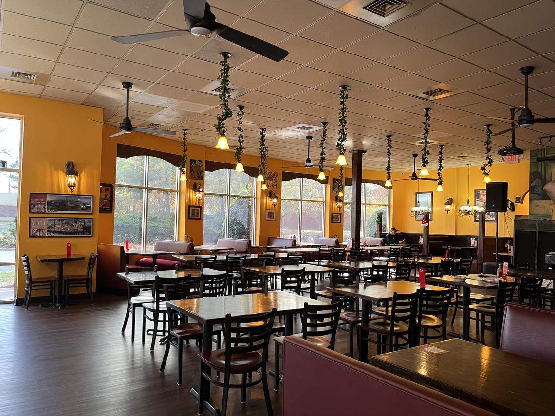 A wide, eye-level shot of an empty restaurant dining room with bright yellow walls and dark wood-look floors. Large arched windows line the back wall, flanked by long reddish-brown booths. The ceiling features several black fans and hanging light fixtures wrapped in decorative ivy.