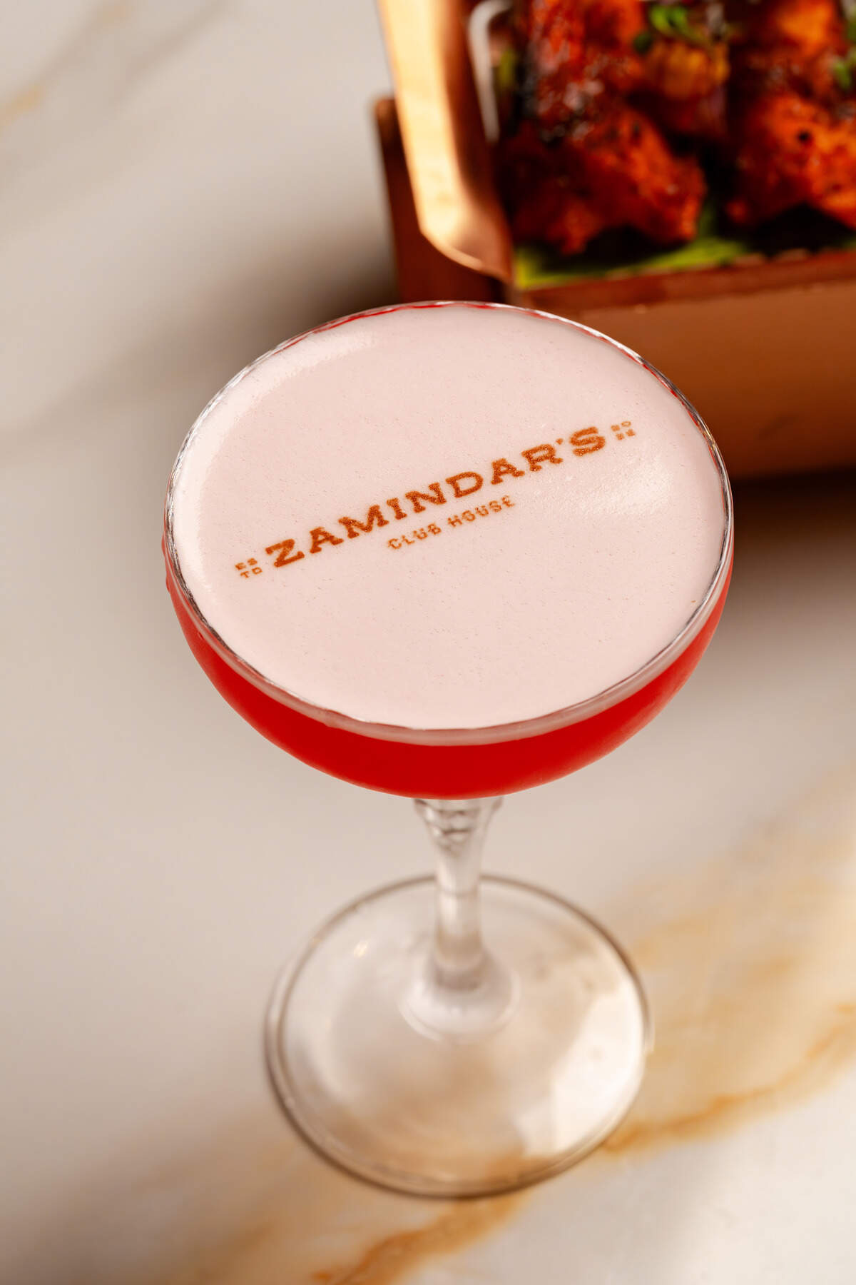 A close-up, high-angle shot captures a bright pink cocktail served in an elegant stemmed coupe glass on a marble surface. The drink features a thick, smooth layer of white foam on top, precisely dusted with cocoa to read “ZAMINDAR’S CLUB HOUSE.” In the blurred background, a warm-toned dish of food sits in a copper serving container.