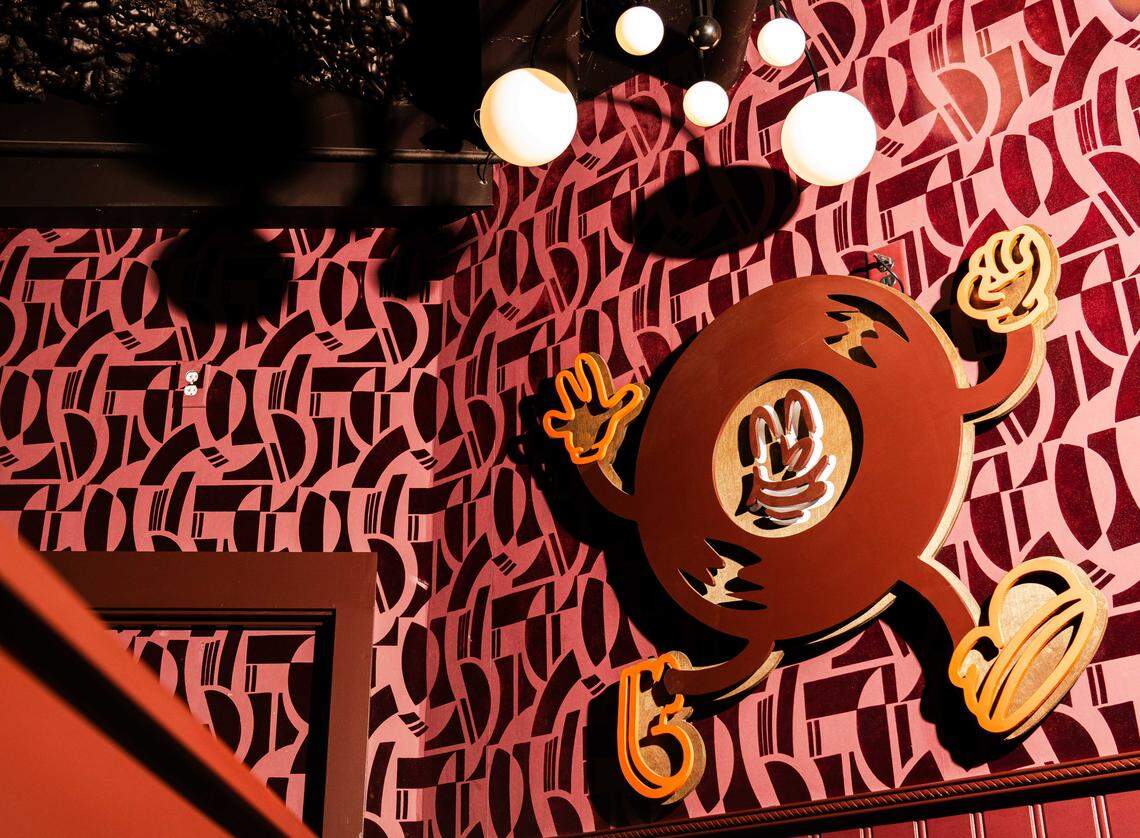 A close-up shot of a corner in the venue featuring deep pink and burgundy geometric-patterned wallpaper. Attached to the wall is a large, stylized 2D figure resembling a brown disc with cartoonish arms and legs in a walking pose. Above the figure, several white globe-style pendant lights hang from a dark, textured ceiling, casting soft light and shadows against the bold wall pattern.