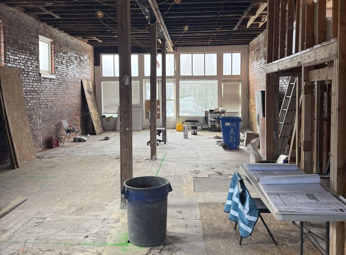 A wide interior view of a historic building under renovation. The space is a long, narrow room with exposed red brick walls, high wooden ceiling joists, and a dusty concrete floor. Large windows at the far end let in bright natural light. A folding table in the foreground holds architectural blueprints and a blue safety vest.