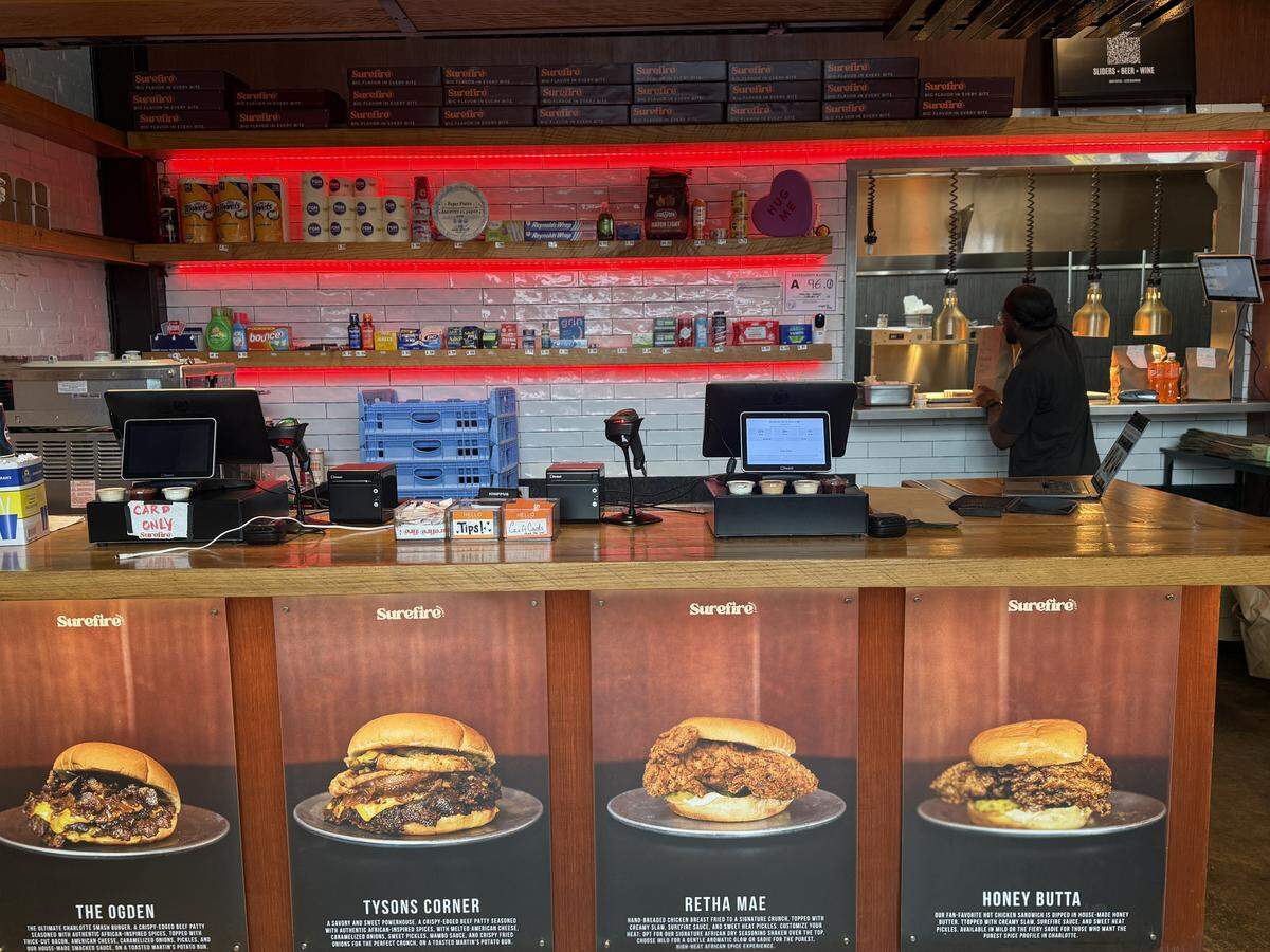 A wide wooden order counter features digital POS systems and menu posters for signature burgers and sandwiches. The front of the counter displays large, backlit photos of menu items: The Ogden, Tysons Corner, Retha Mae, and Honey Butta.