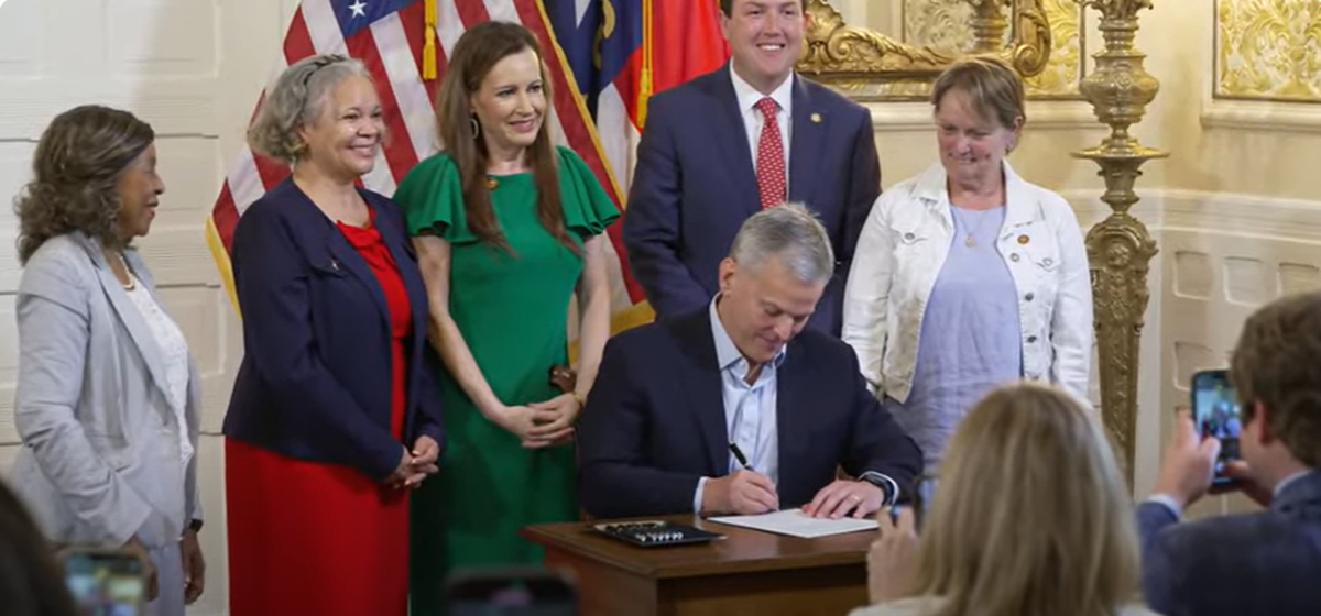 NC Gov. Josh Stein, flanked by Charlotte-area leaders, signs a bill allowing Mecklenburg County to put a sales tax referendum on the ballot to pay for transportation projects. Behind Stein is Charlotte Mayor Vi Lyles (in red dress) and State Rep. Tricia Cotham (in green dress).