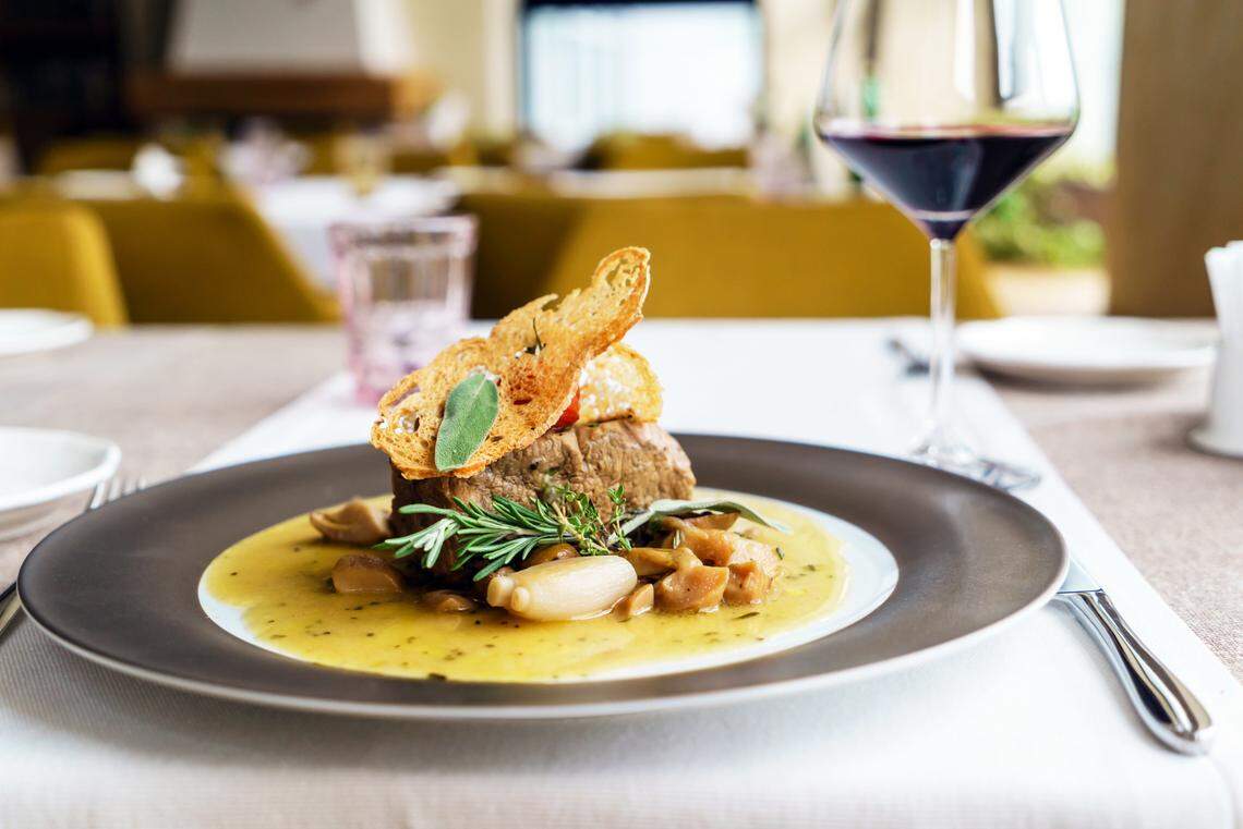 A gourmet beef entrée plated on a dark rimmed dish. The sliced beef rests in a savory yellow sauce with roasted mushrooms, a pearl onion, and sprigs of fresh rosemary and sage. Two thin, crispy crostini crackers are perched on top of the meat. A glass of red wine stands in the blurred background of the elegantly set dining room.