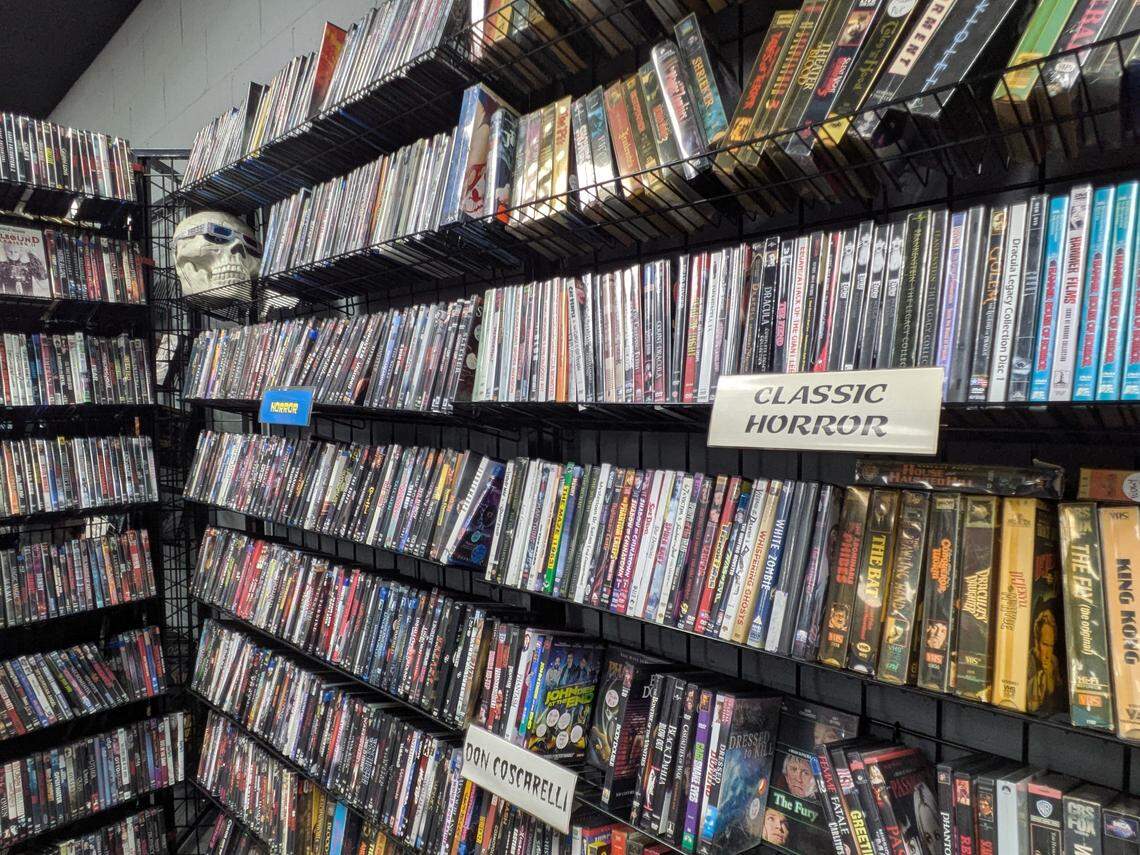 A wide-angle shot looking into the corner of a video store or large personal collection, featuring floor-to-ceiling black wire shelves packed with hundreds of DVDs. A white sign reading “CLASSIC HORROR” is visible, with box sets for films like “King Kong” and “White Zombie” on the shelf below. A white skull wearing sunglasses is perched on an upper shelf to the left.