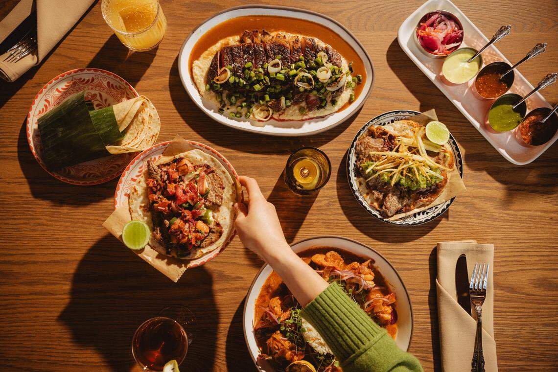 An overhead “flat lay” photo of a full Mexican feast on a warm wooden table. The spread includes a variety of colorful dishes including a large plate of sliced steak topped with charred green onions; a bowl of shrimp and rice in red sauce; and two different tacos on patterned plates. Also shown are a side of warm tortillas wrapped in a banana leaf and a long white platter holding five small bowls of colorful salsas and pickled onions. A hand in a green sweater reaches into the frame toward a taco, adding a sense of communal dining.