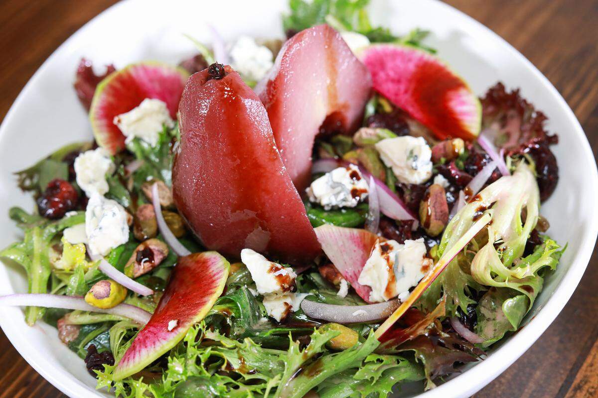 A vibrant gourmet salad served in a white bowl, centered around a halved pear poached to a deep ruby red. The salad features a mix of leafy greens, including frisée, topped with thinly sliced watermelon radishes showing their signature pink centers and green rims. It is garnished with crumbles of blue cheese, whole roasted pistachios, dried cranberries, and thin slivers of red onion. A dark balsamic glaze or wine reduction is drizzled over the ingredients, highlighting the textures of the fruit and nuts.