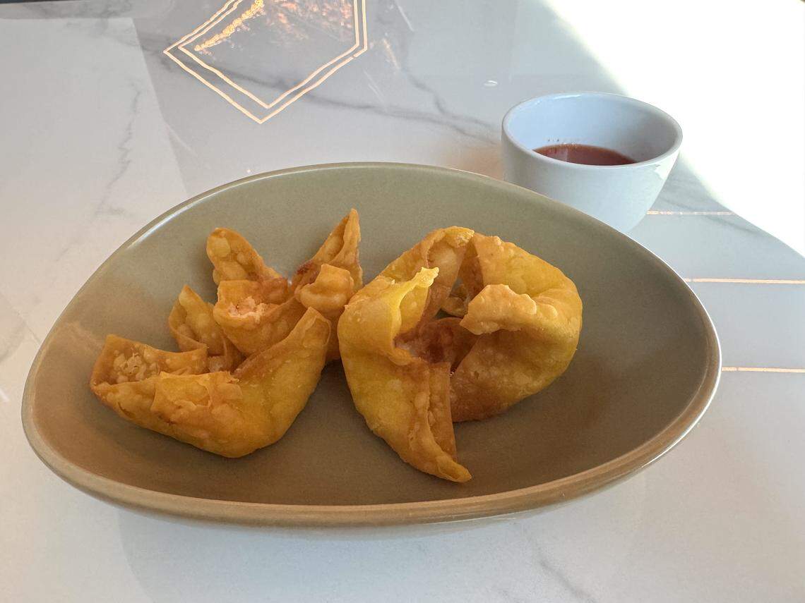 A close-up, high-angle shot of three golden-brown fried wontons served in a shallow, matte-green oval bowl. A small white ceramic cup of red dipping sauce sits in the background on a white marble-patterned tabletop.
