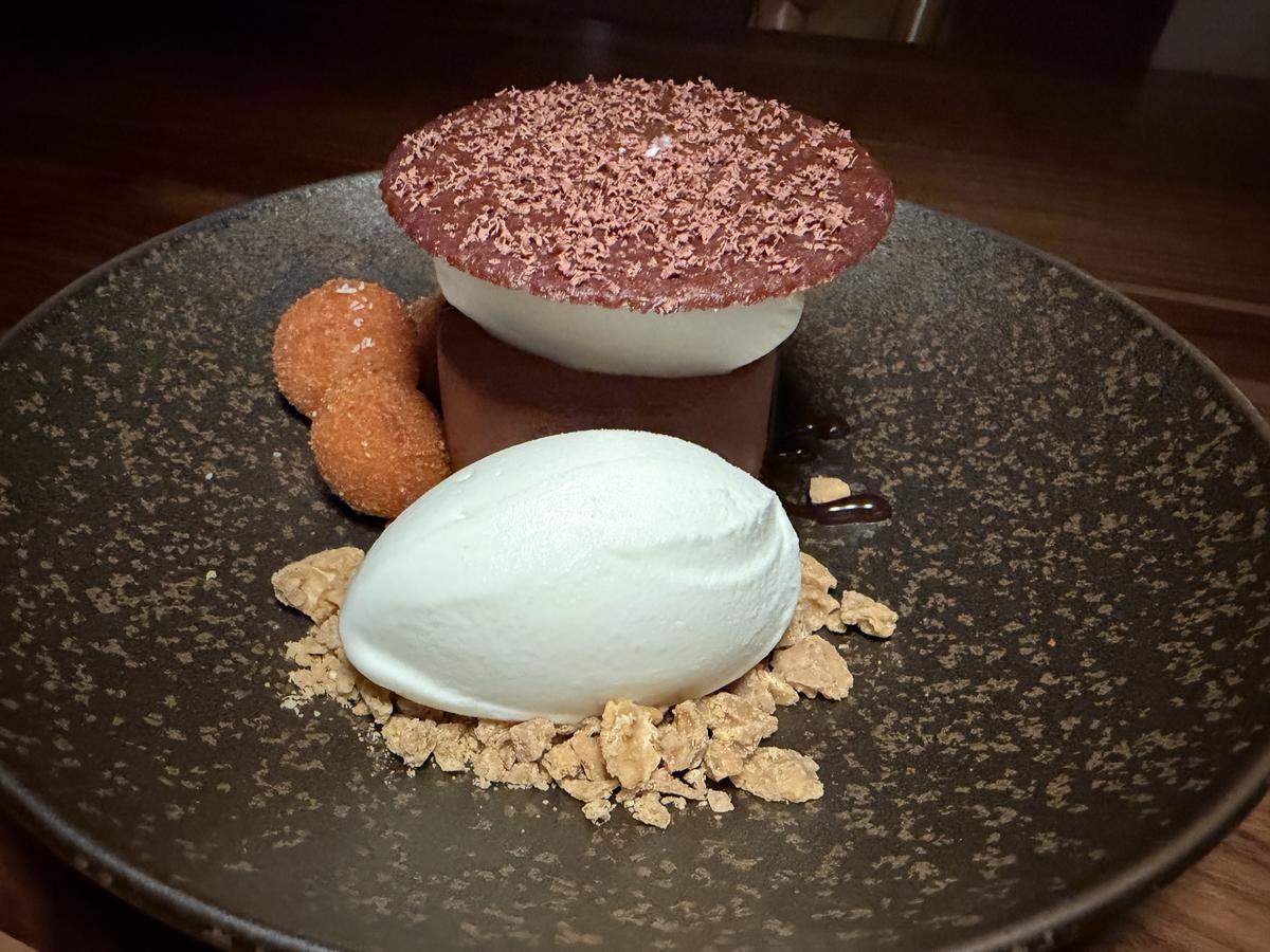 A gourmet, deconstructed chocolate dessert is presented on a dark, textured stoneware plate. The main element is a vertical stack with a thick, cylindrical ring of dark chocolate ganache or dense chocolate mousse, a layer of smooth, white chantilly cream or milk-based mousse and a thin, delicate chocolate tuile or wafer, heavily dusted with fine cocoa powder or chocolate shavings.