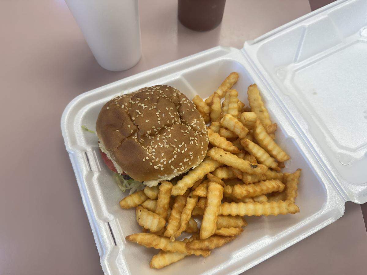 A high-angle, close-up shot of a burger and crinkle-cut fries served in an open white styrofoam to-go container. The burger is on a sesame seed bun, and the golden-brown fries are piled next to it. The container rests on a light-colored table, with a white styrofoam cup partially visible in the top left.