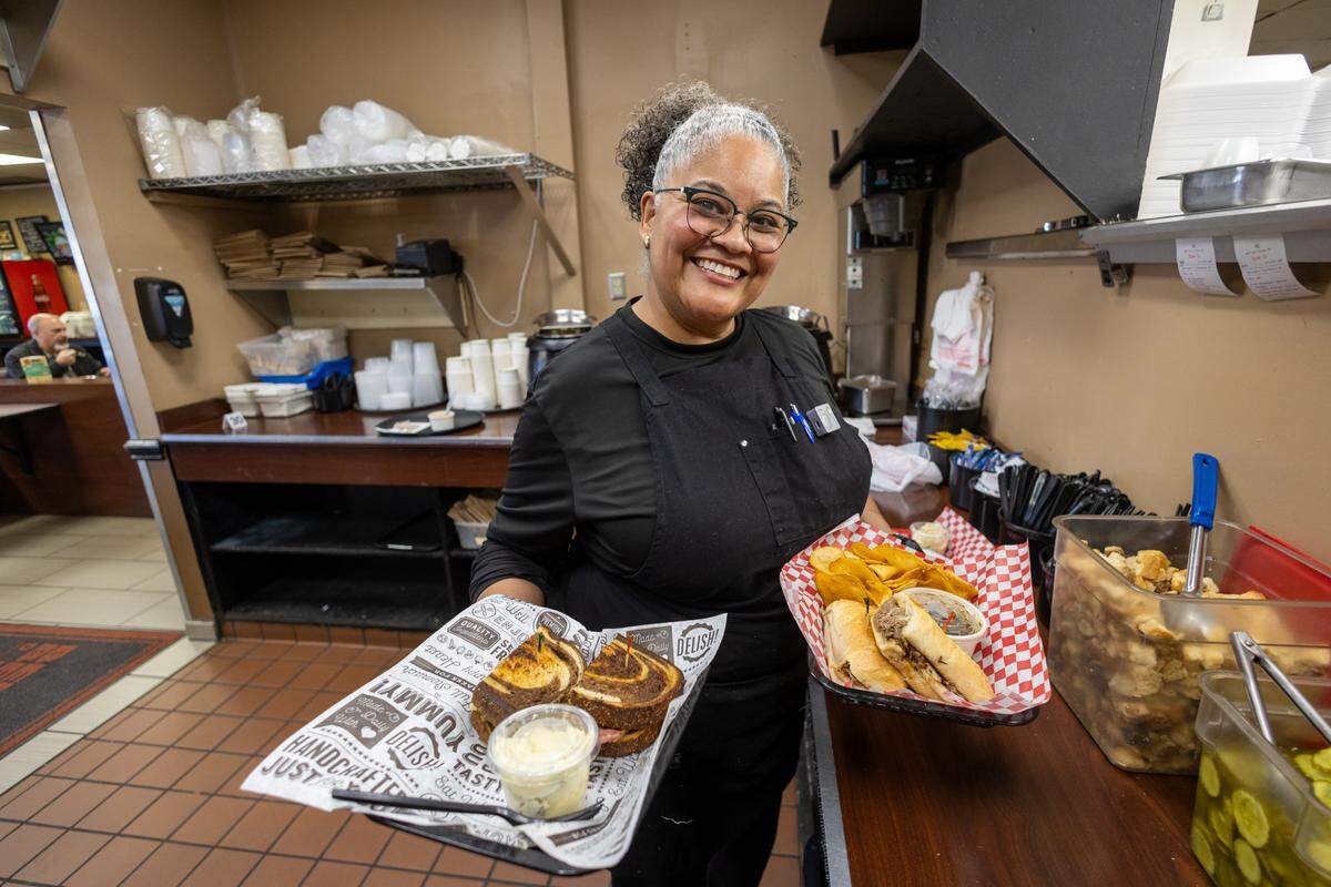 A smiling person in a black chef’s coat and glasses stands in a kitchen prep area, holding two black trays. One features a toasted marble rye sandwich with a side of potato salad, and the other holds a hot sub sandwich with a side of potato chips.