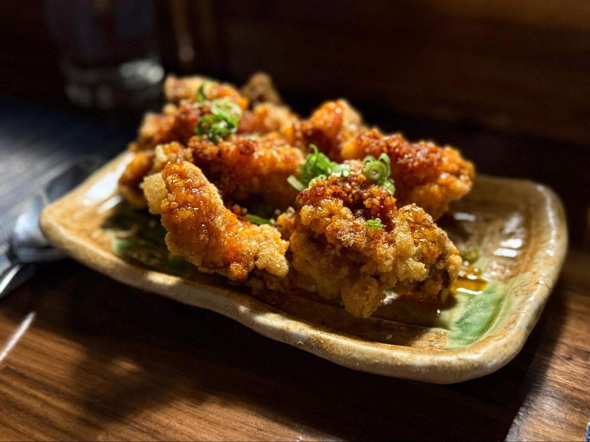 A close-up, shallow-depth-of-field shot of crispy fried chicken pieces glazed in a reddish-orange spicy sauce. The chicken is served on a rustic tan ceramic plate and garnished with fresh green scallion rings.