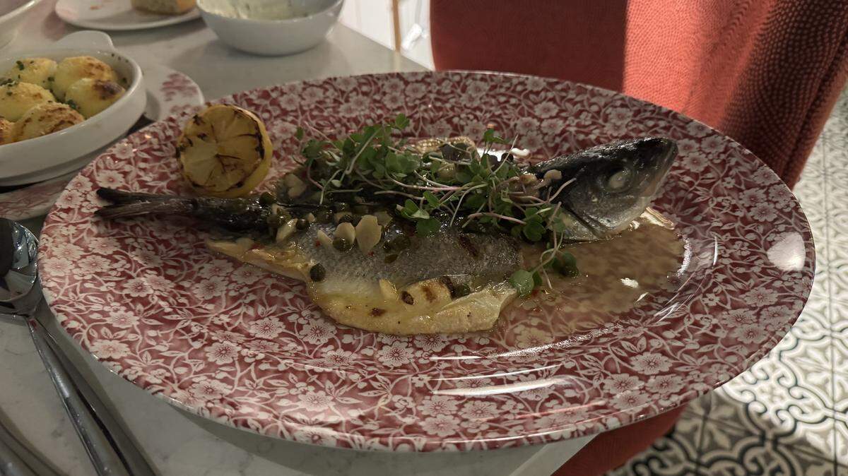 A whole roasted or grilled fish served in a shallow pool of light sauce on a red-and-white floral patterned oval platter. The fish is presented head-on, topped with capers, slivered garlic, and fresh green microgreens. A charred lemon half rests on the side of the plate.