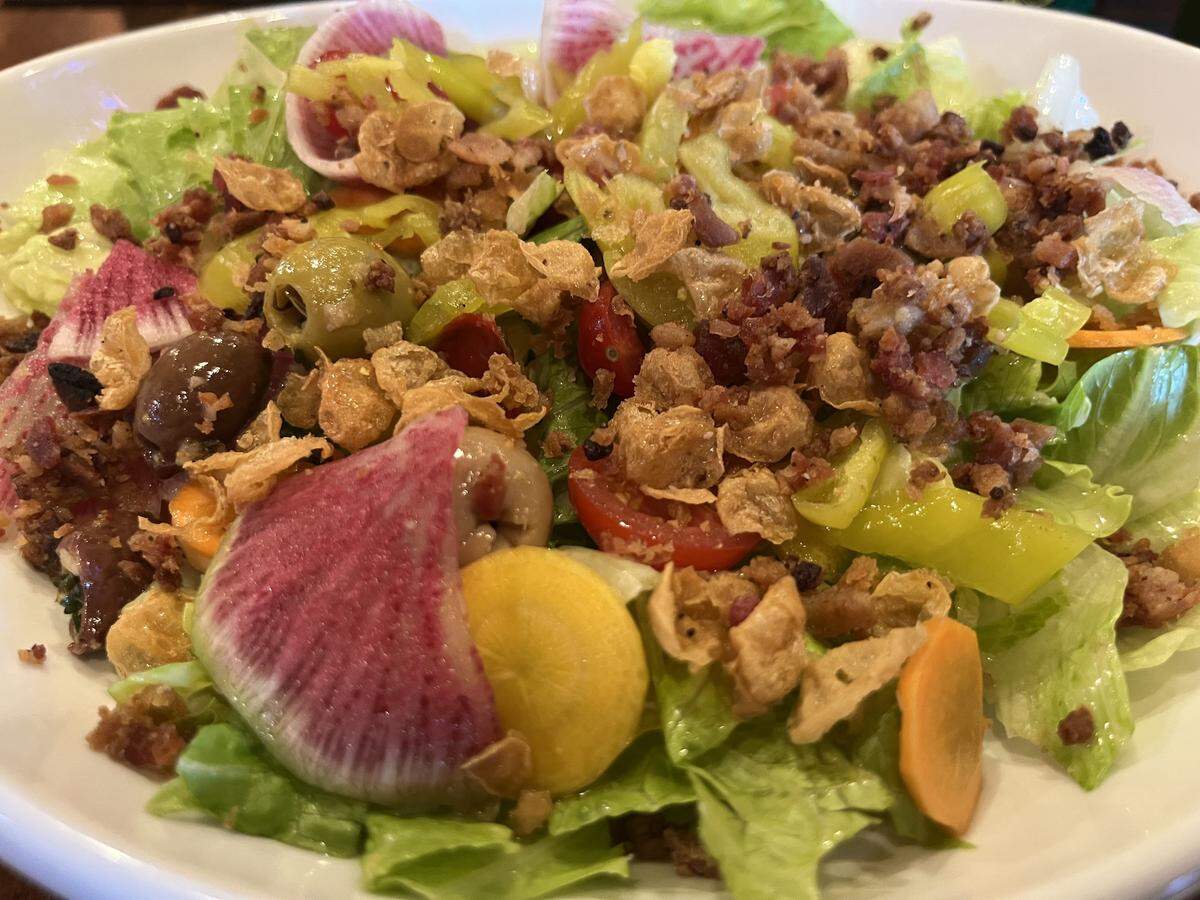 A detailed, close-up photo of a restaurant salad. The bowl is filled with fresh lettuce and a generous amount of toppings, including crumbled bacon, bright yellow pepperoncini, dark olives, red tomatoes, and decorative slices of watermelon radish. The salad is finished with a sprinkle of crispy fried cannellini beans