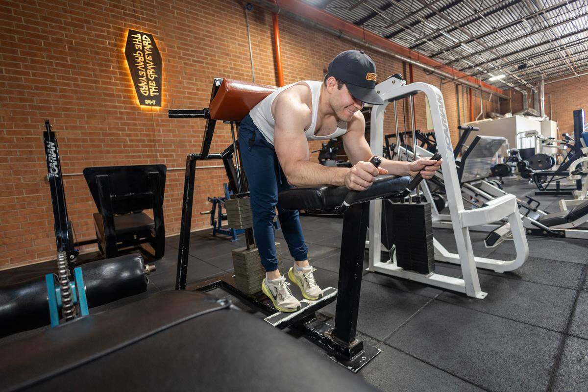 Gym user in a white tank top and dark baseball cap using a black standing vertical press machine. A red brick wall and neon coffin sign are visible in the background.
