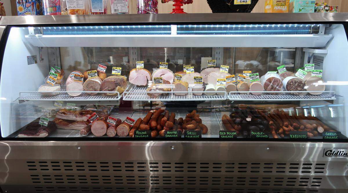 A brightly lit, stainless steel deli case filled with a variety of meats and sausages. Labels identify different types, including Polish sausage, kabanos, and garlic sausage, along with various sliced cold cuts on the top shelf.