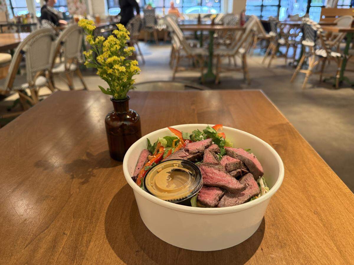 The salad is served in a white, disposable bowl and is topped with several slices of medium-rare grilled steak. Visible salad components include mixed greens, sliced red bell peppers, and what appear to be fresh herbs like cilantro. A small, clear plastic container of tan/yellow dressing sits in the center of the steak. To the left of the bowl is a small, dark amber glass vase holding a few sprigs of small yellow flowers. The background is blurred, showing the interior of a restaurant with tables featuring wooden tops and chairs with white woven seats and dark metal frames. The lighting suggests a daytime setting.