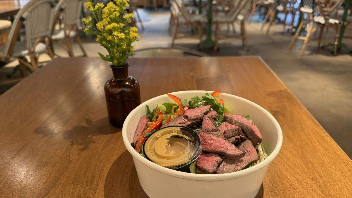 The salad is served in a white, disposable bowl and is topped with several slices of medium-rare grilled steak. Visible salad components include mixed greens, sliced red bell peppers, and what appear to be fresh herbs like cilantro. A small, clear plastic container of tan/yellow dressing sits in the center of the steak. To the left of the bowl is a small, dark amber glass vase holding a few sprigs of small yellow flowers. The background is blurred, showing the interior of a restaurant with tables featuring wooden tops and chairs with white woven seats and dark metal frames. The lighting suggests a daytime setting.